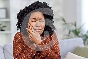 African american young woman sitting on sofa at home and holding hands to cheek, eyes closed, suffering from toothache