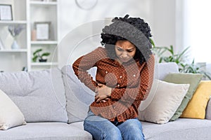 African American young woman sitting on sofa at home and holding her side of body and stomach. feels severe pain, needs