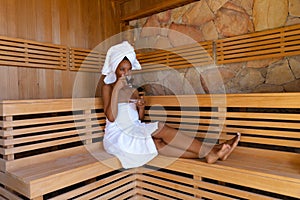 African american young woman wearing bathrobe drinking tea while sitting on wooden bench at sauna