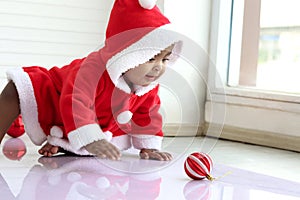 African baby kid in red Santa Claus costume crawling to grab ball ornament in white living room, beautiful little child girl