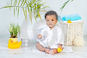 an African baby in a white coat after a bath and bathing plays with a duck at home, the concept of care and hygiene of