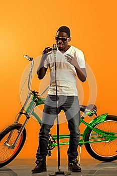 African black man singing at microphone with a bicycle in back on orange background