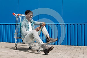 African black man sitting on a shopping cart with a phone in his hands, shopping online