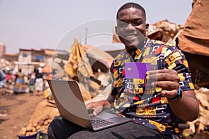 African boy shows his credit card while using his laptop, he is at the market. Credit card payments in Africa concept