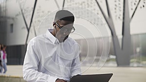 African Businessman with headphones Sitting on bench in park and working on notebook