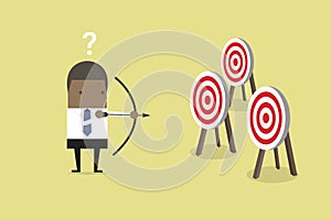 African businessman holding bow and arrow confused by multiple bulls eye target.