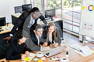 African businessman stands in the back of Caucasian colleagues looking on laptop at an internal business group meeting.