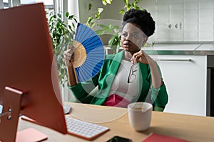 African businesswoman uses hand fan sits at desk with computer in summer day, suffers from heat.