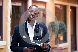 African doctor with stethoscope wearing classic suit reading a book  near modern clinic hospital building