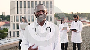 African doctor in white lab coat posing on camera with stethoscope and smiling.