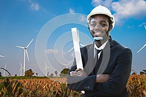 African engineer wearing white hard hat standing with against wind turbine on sunny day