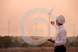 African engineer wearing white hard hat standing with digital tablet against wind turbine