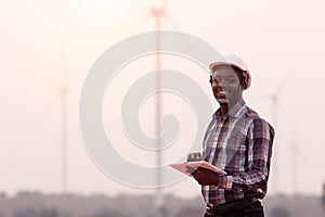 African engineer wearing white hard hat standing with digital tablet against wind turbine on sunny day