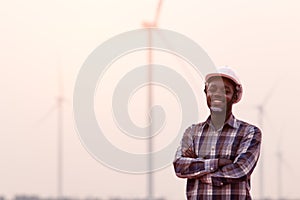 African engineer wearing white hard hat standing with digital tablet against wind turbine on sunny day