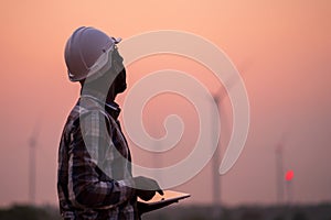 African engineer wearing white hard hat standing with digital tablet against wind turbine on sunny day