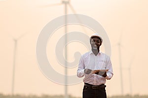 African engineer wearing white hard hat standing with digital tablet against wind turbine on sunny day