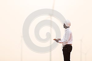 African engineer wearing white hard hat standing with digital tablet against wind turbine on sunny day