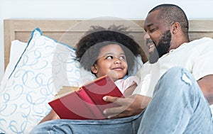 African family, single father and cute boy, lying on bed, reading a book together at home. Beard dad spends time and have fun