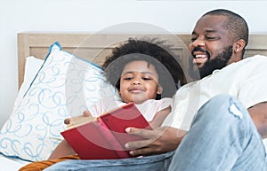 African family, single father and cute boy, lying on bed, reading a book together at home. Smiling beard dad spends time and have