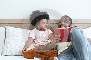 African family, single father and cute boy, lying on bed, reading a book together at home. Smiling beard dad spends time and have