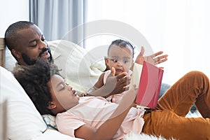 African family, single father, cute newborn baby and boy, lying on bed, reading a book together at home. Beard dad spends time,