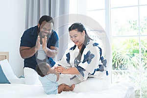 African father and Asian mother cheer up cute newborn baby playing sit up exercise on bed, smiling looking at innocent infant with