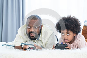 African father and child son playing online video games, using joysticks or game console, having fun at home while lying on bed