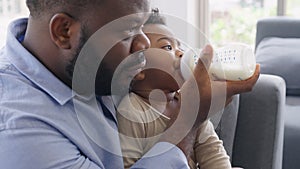 African father Sitting on sofa, holding and feeding newborn baby from bottle milk in living room. Authentic lifestyle candid real
