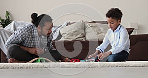 African father and son play toy railroad seated on carpet