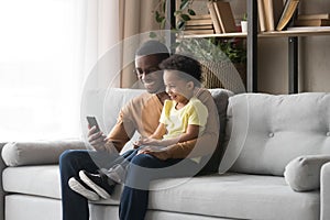 African father son sitting on couch having fun use smartphone