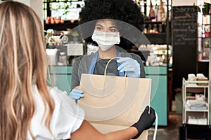 African female cafe worker wear face mask giving takeaway food bag to customer.