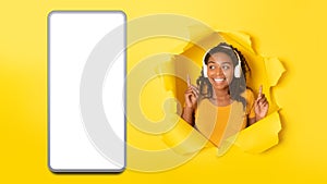 African Female In Headphones Near Big Smartphone Screen, Yellow Background