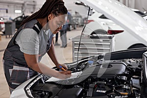 african Female mechanic at work, checking car hood writing notes in tablet