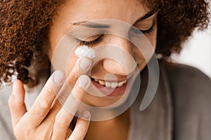 African girl applying face moisturizer cream close-up to protect skin from dryness in bath. African american woman in