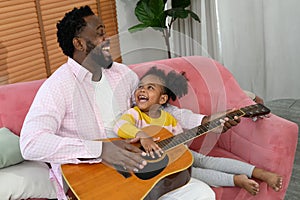 African girl and father are playing guitar and smiling while sitting on couch