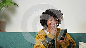 African girl holding smartphone touch screen typing scroll page at home. Woman with cell phone surfing internet using
