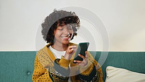 African girl holding smartphone touch screen typing scroll page at home. Woman with cell phone surfing internet using