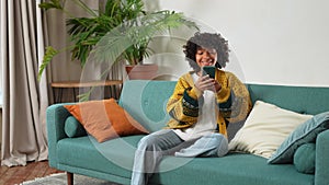 African girl holding smartphone touch screen typing scroll page at home. Woman with cell phone surfing internet using