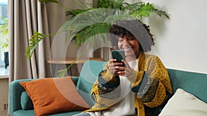 African girl holding smartphone touch screen typing scroll page at home. Woman with cell phone surfing internet using