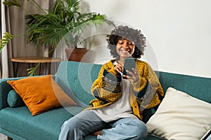 African girl holding smartphone touch screen typing scroll page at home. Woman with cell phone surfing internet using