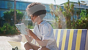 African girl reading book sitting on street bench close up. Student studying