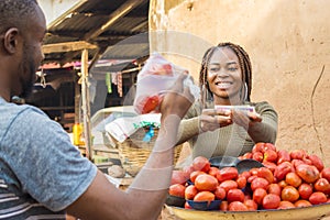 african girl selling tomatoes in a local african market to a male customer smiling while receiving payment