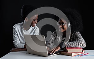 African girl wear glasses and headphones smiling at little brother while they are online learning with laptop textbook