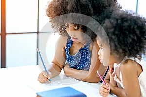 African girls as older and younger sister write or draw something on white paper near the book in front of glass windows with day