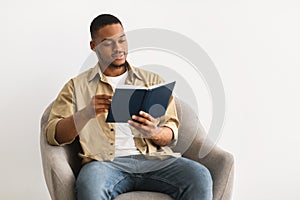 African Guy Reading Book Sitting In Chair Over Gray Background