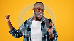 African Guy Singing Holding Phone Like Microphone Over Yellow Background