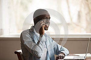 African guy sit at table near computer talk on smartphone
