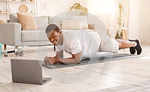 African Guy Training At Laptop Doing Plank Exercise In Bedroom