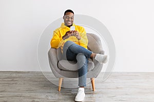 African Guy Using Smartphone Sitting In Chair Over Gray Wall