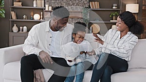 African happy family parents with child daughter sitting on sofa at home having fun sincerely rejoice child with teddy